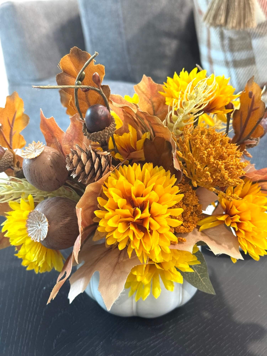 back side of lush elegant handmade small fall arrangement with yellow and gold flowers, brown leaves, poppy heads, acorns, pinecones in white ceramic pumpkin shaped vase styled on coffee table