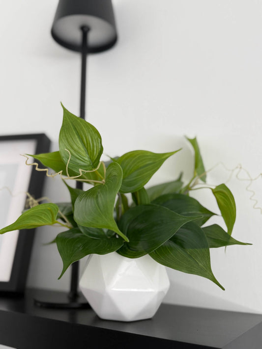 small realistic faux plant of green leaves in white ceramic pot vase of geometric shape styled on shelf