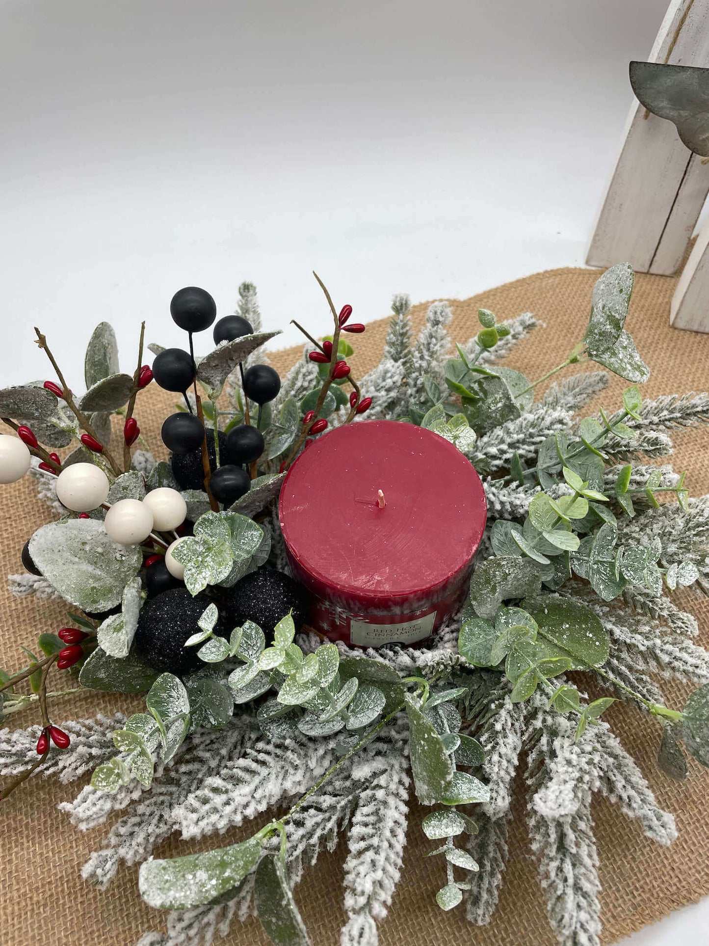 small table wreath with frosted evergreen and greenery stems, red, white and black ornaments and berries, and red pillar candle in the middle