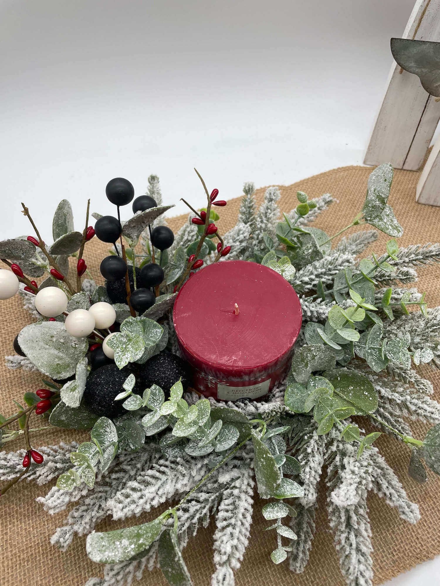 small table wreath with frosted evergreen and greenery stems, red, white and black ornaments and berries, and red pillar candle in the middle