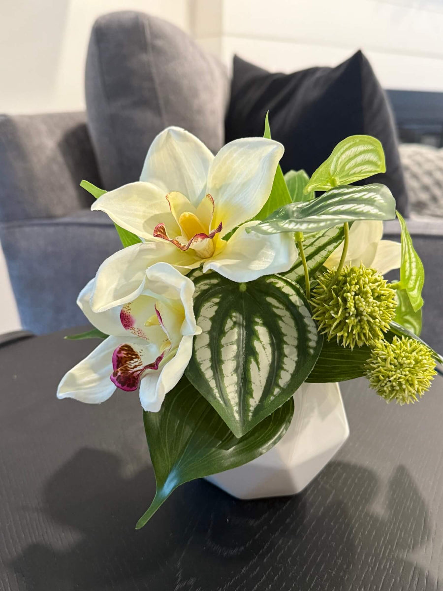small realistic looking white orchid arrangement with greenery in white ceramic pot vase of geometric shape set on coffee table