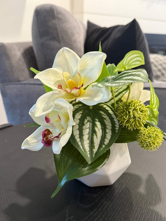 small realistic looking white orchid arrangement with greenery in white ceramic pot vase of geometric shape set on coffee table