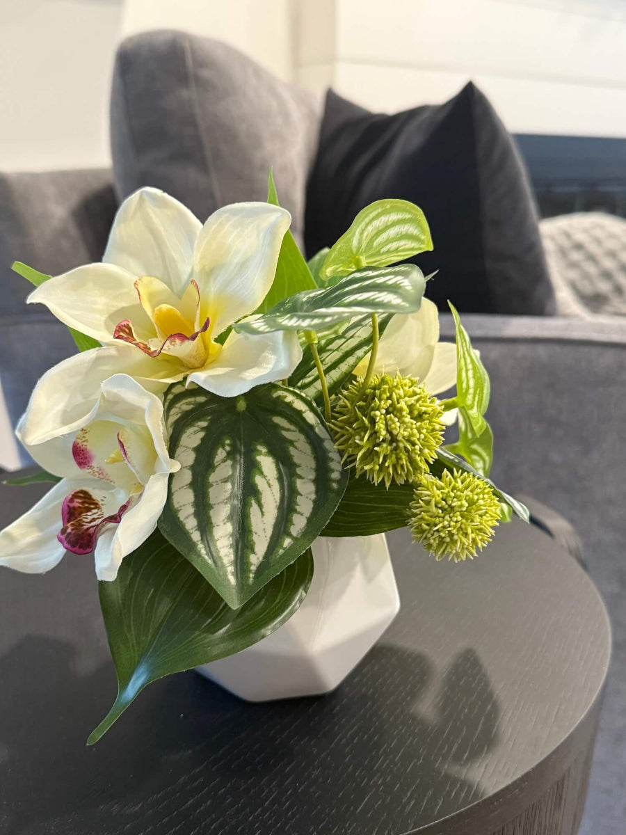 small realistic looking white orchid arrangement with greenery in white ceramic pot vase of geometric shape set on coffee table