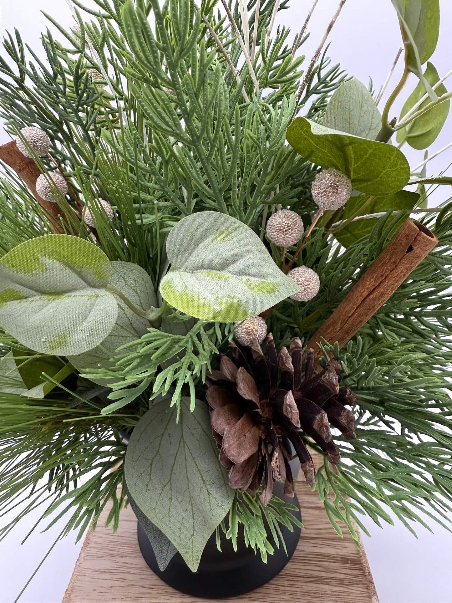 back of winter arrangement with variety of evergreen stems in black ceramic vase with cinnamon sticks, pinecones and berries
