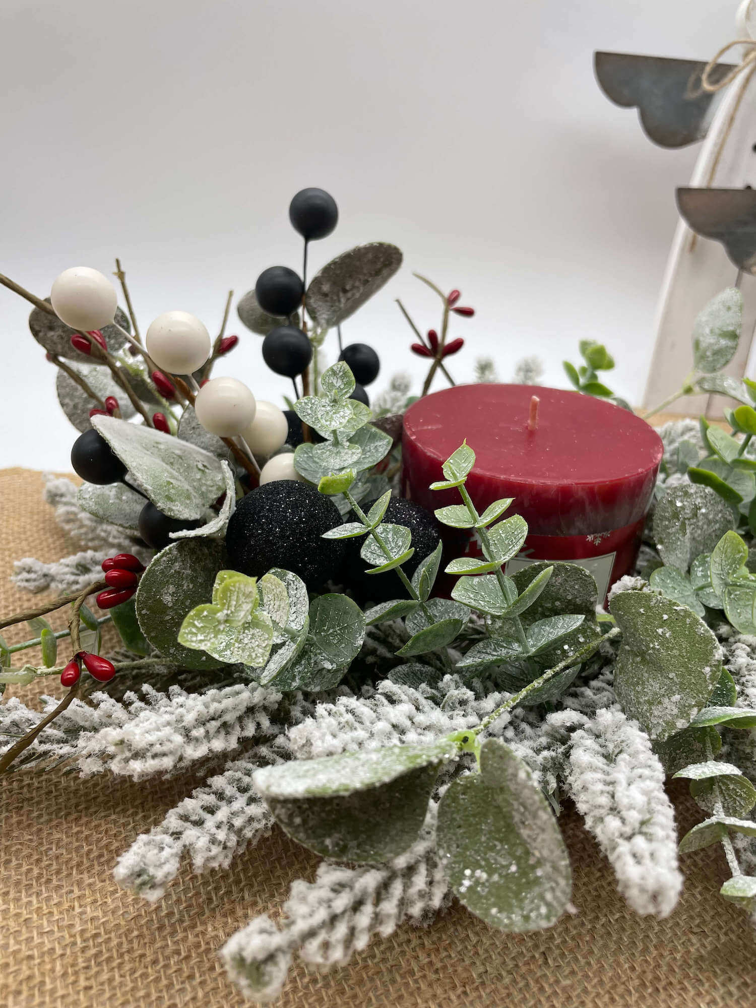 close up for small table wreath with frosted evergreen and eucalyptus stems, red, white and black ornaments and red pillar candle in the middle
