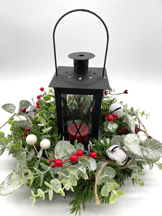 winter evergreen table wreath with frosted greenery, white metal bells, red and white berries and black metal lantern in the middle 