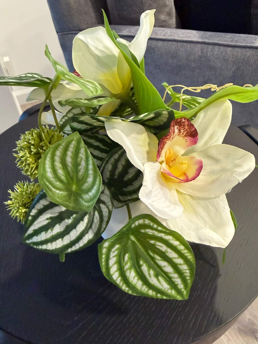 back view of small realistic looking white orchid arrangement with greenery in white ceramic pot vase of geometric shape set on coffee table