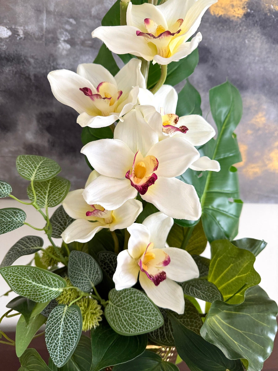 realistic looking white orchid centerpiece with variety of greenery in tall gold ceramic vase