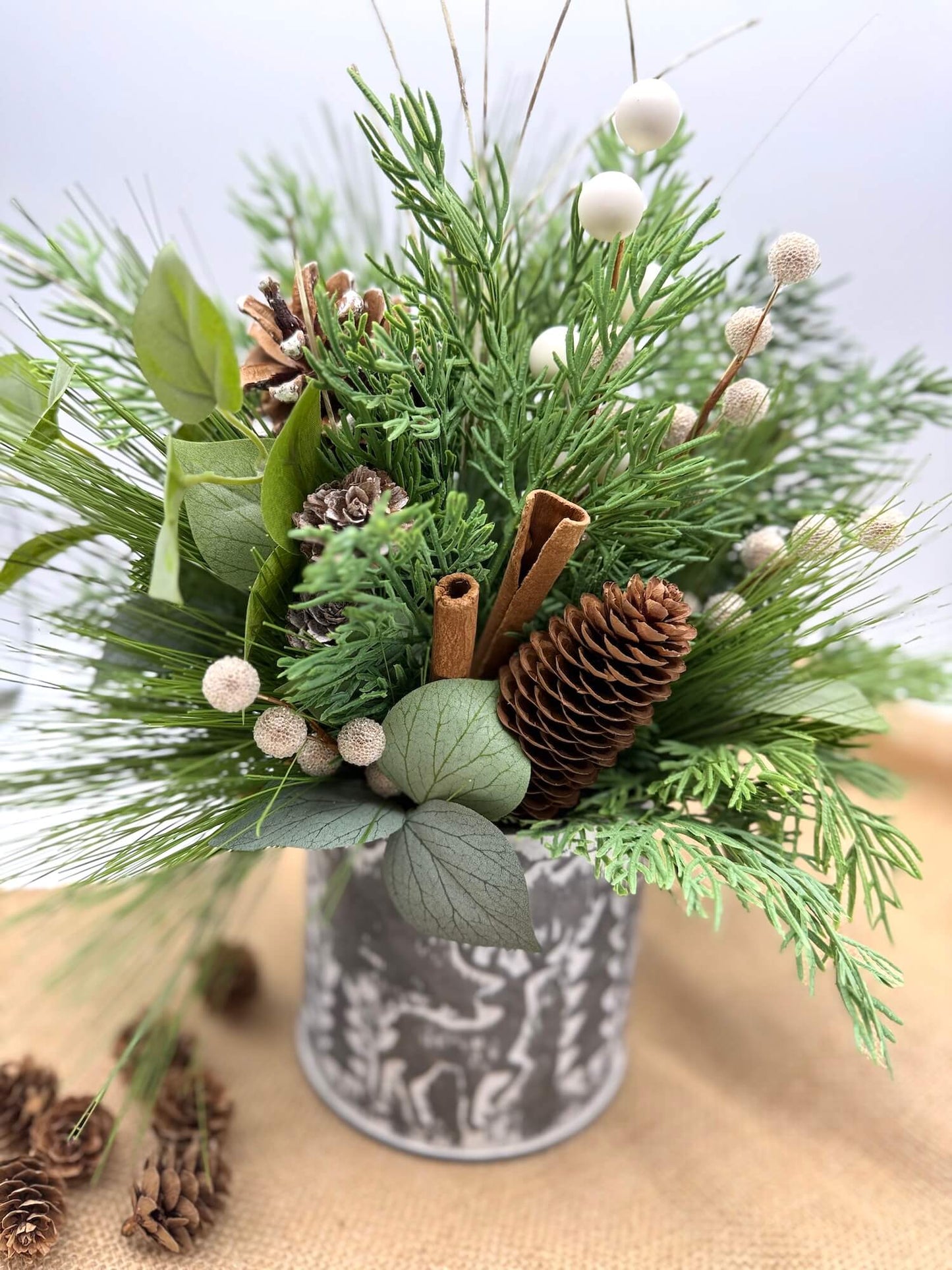 winter arrangement with evergreen and greenery stems, pinecones, cinnamon sticks and white and goldish accents in galvanized tin with forest theme