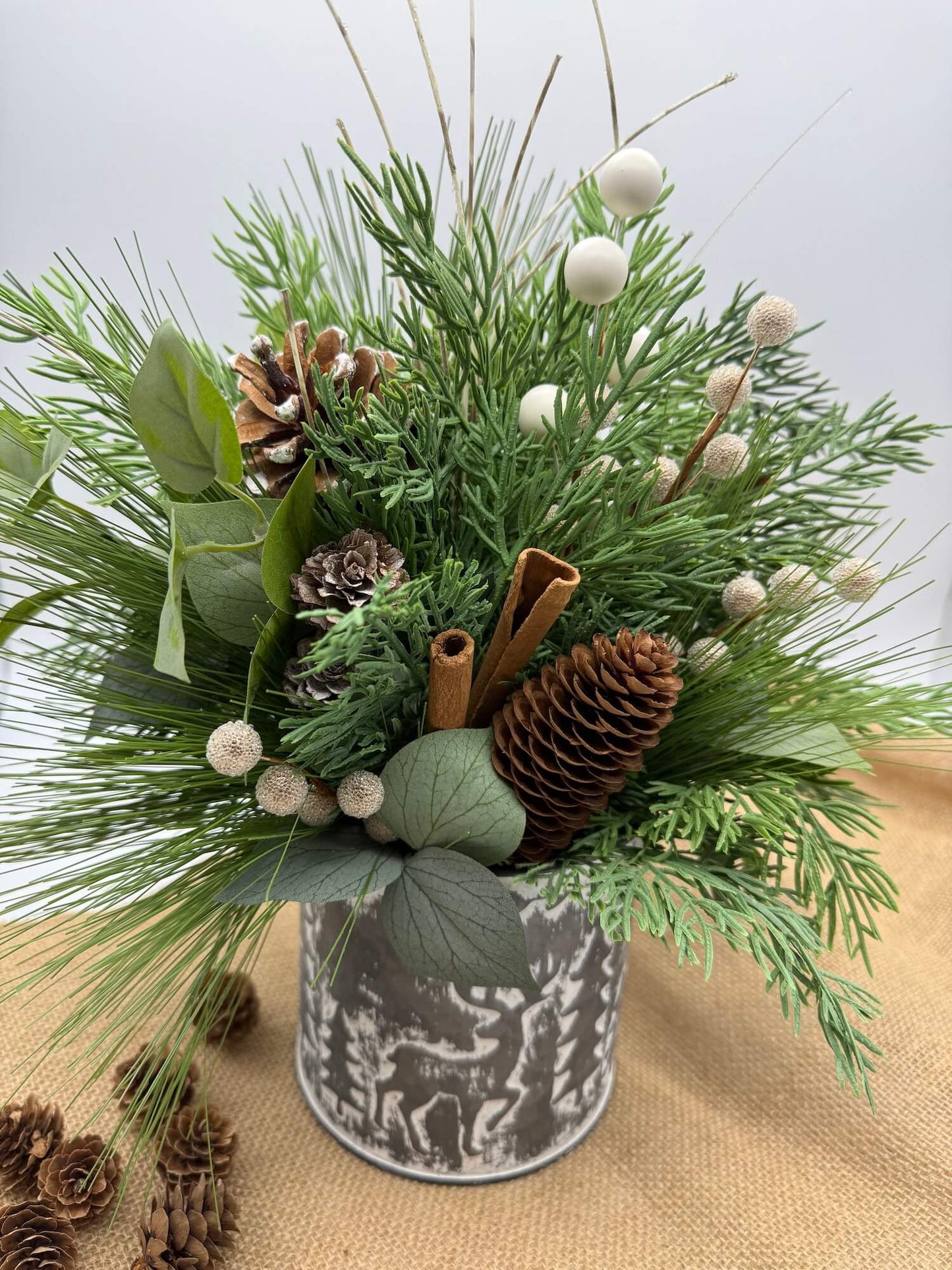 winter arrangement with greenery, evergreen, pinecones, cinnamon sticks, white and gold-ish accents in decorative metal silver-like tin