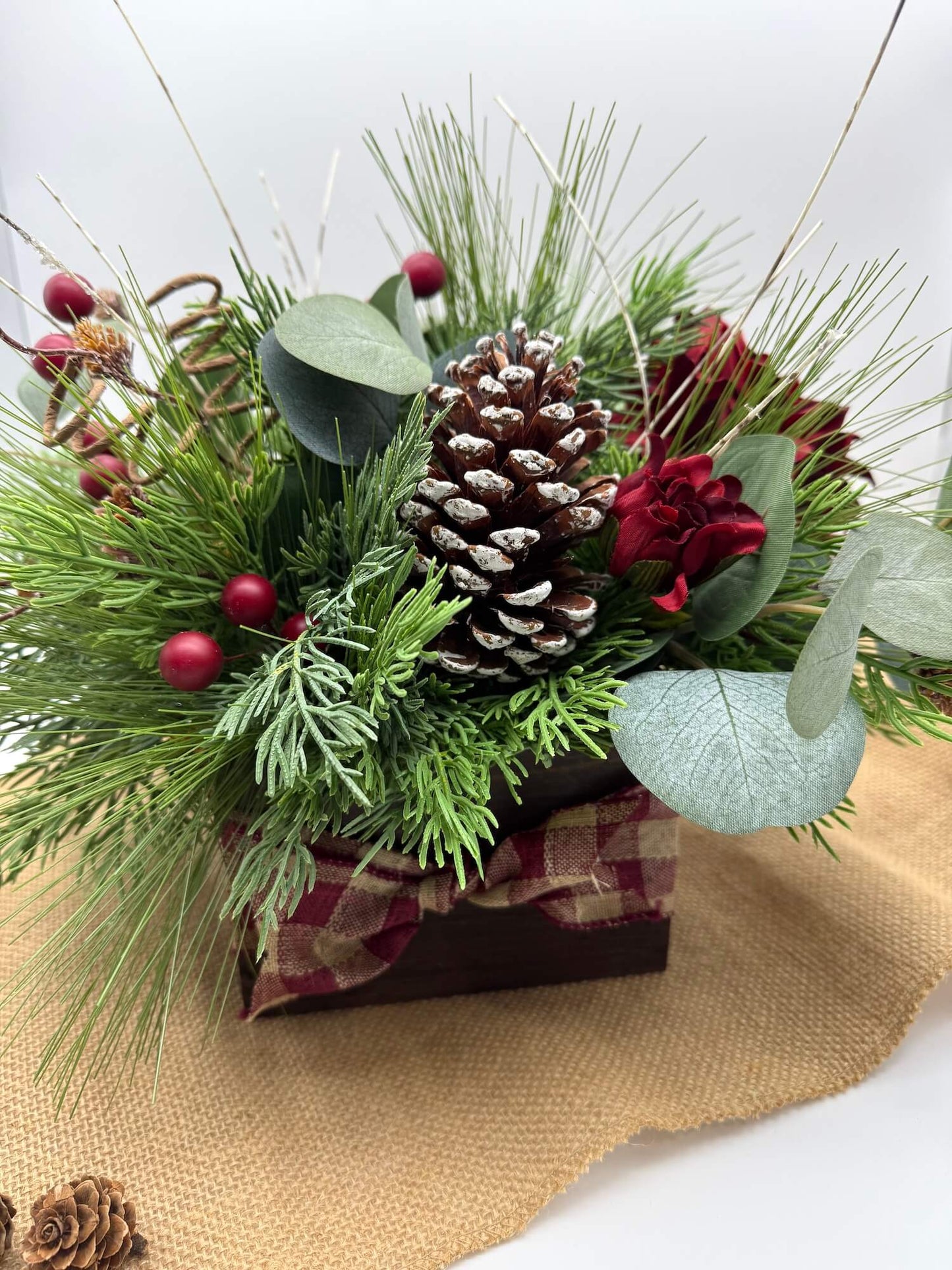 Christmas-themed floral arrangement with pine cones, berries, and greenery in wooden vase with burlap ribbon