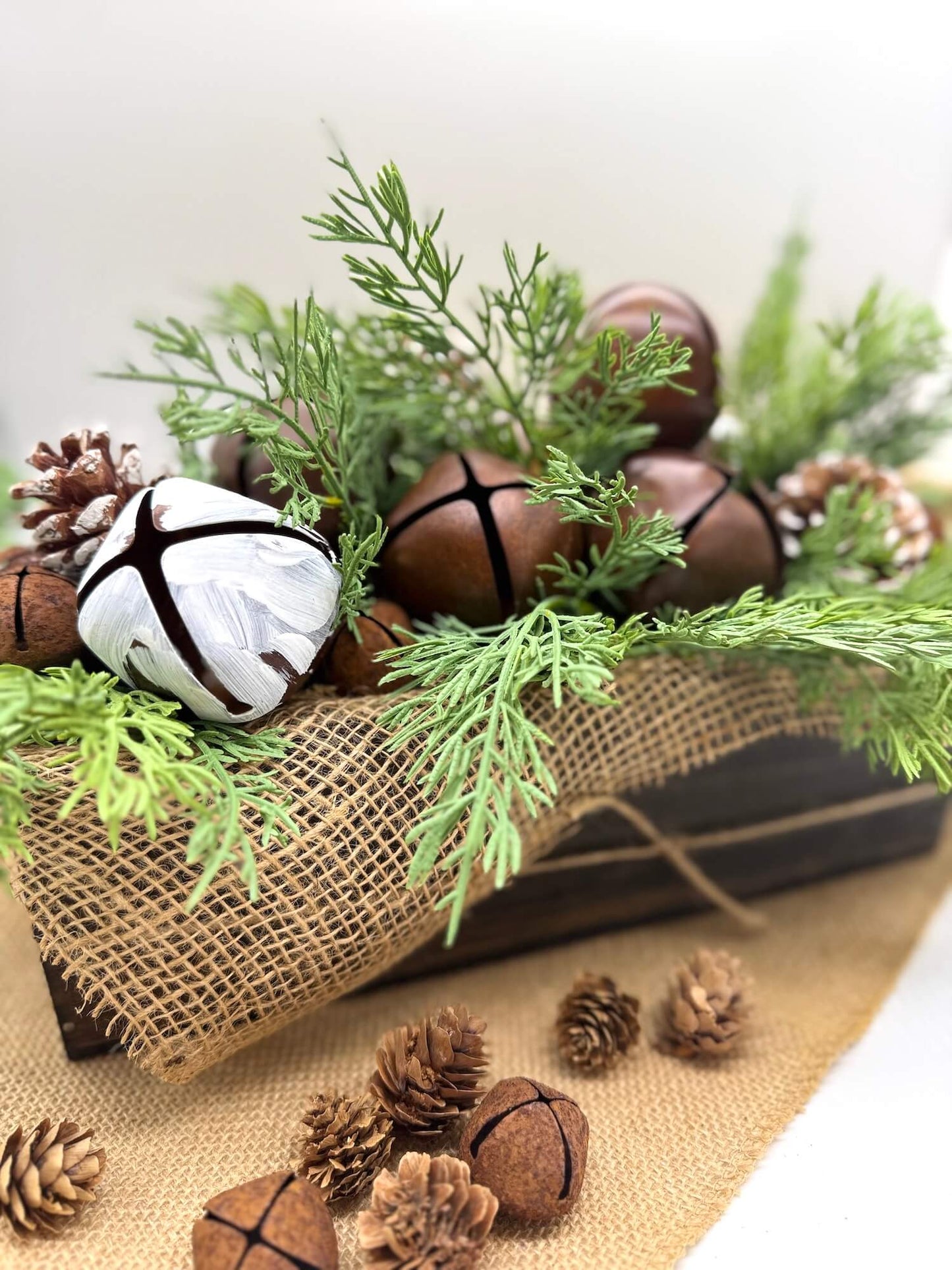 rustic winter arrangement with metal bells in brown and white, pinecones and lush evergreen in wooden planter decorated with burlap