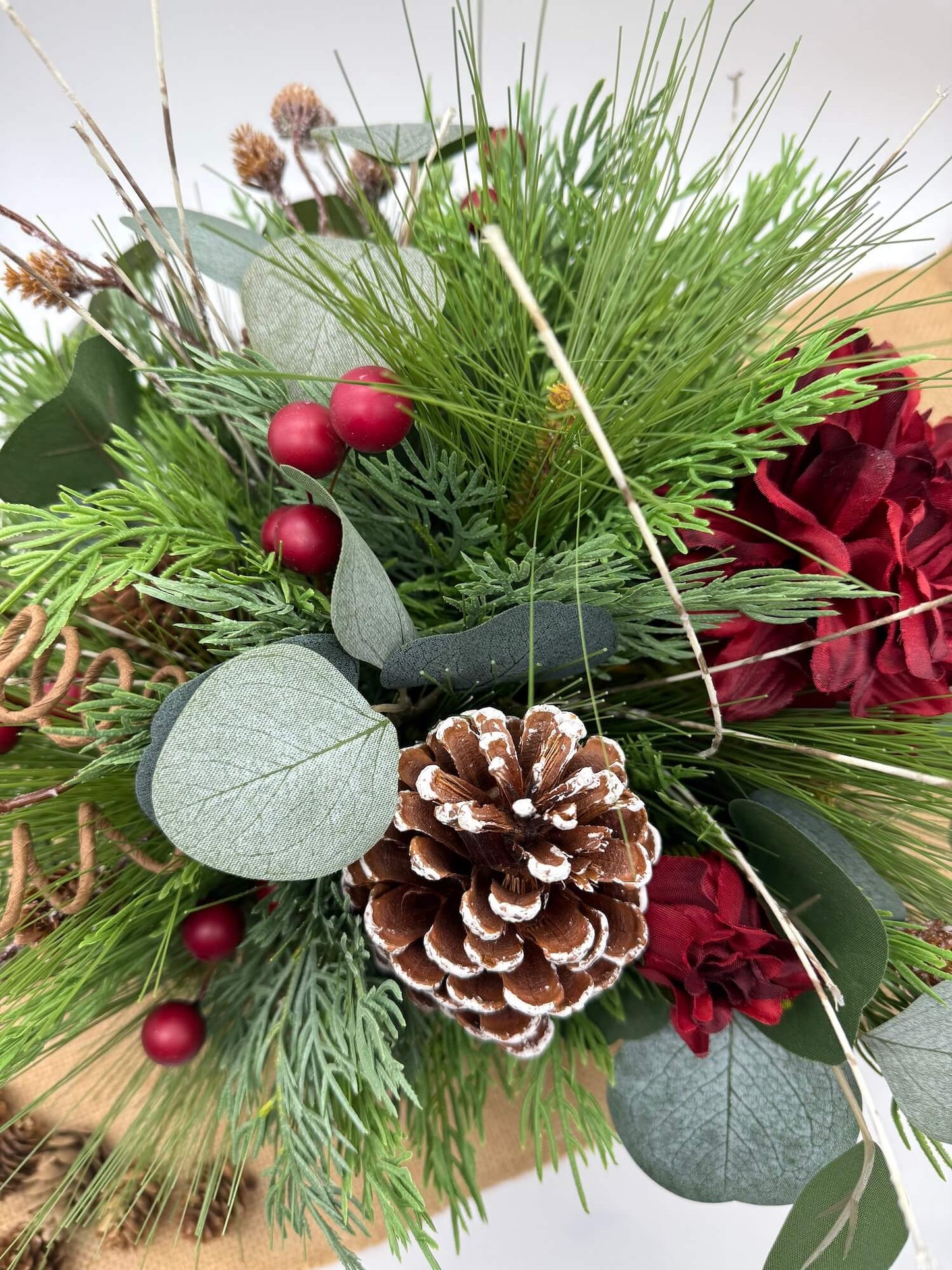 Christmas-themed floral arrangement with greenery, red berries, and pine cones in square wooden vase