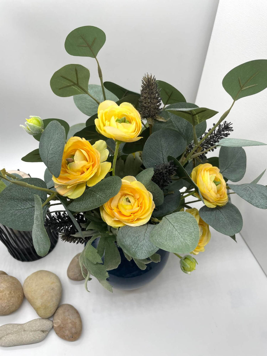 view from top of realistic faux bouquet of yellow runcunculas flowers silver eucalyptus stem and black wheat stems in navy blue ceramic vase