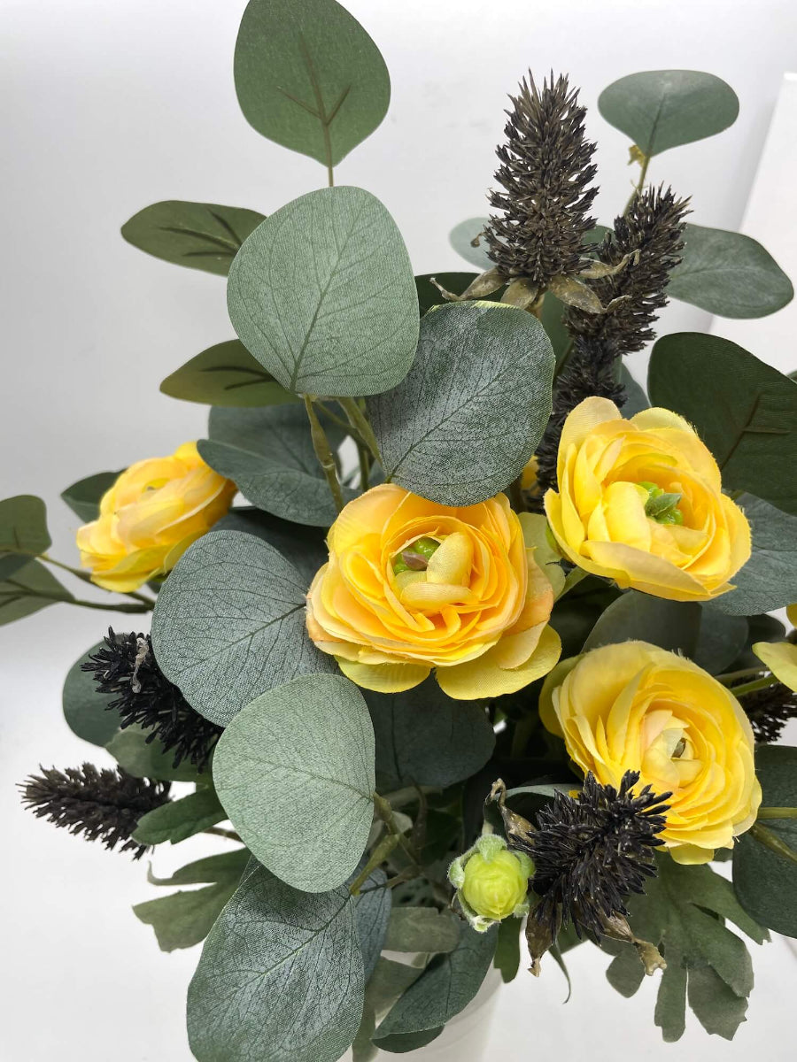 close up for realistic faux bouquet of yellow runcunculas flowers silver eucalyptus stem and black wheat stems in navy blue ceramic vase